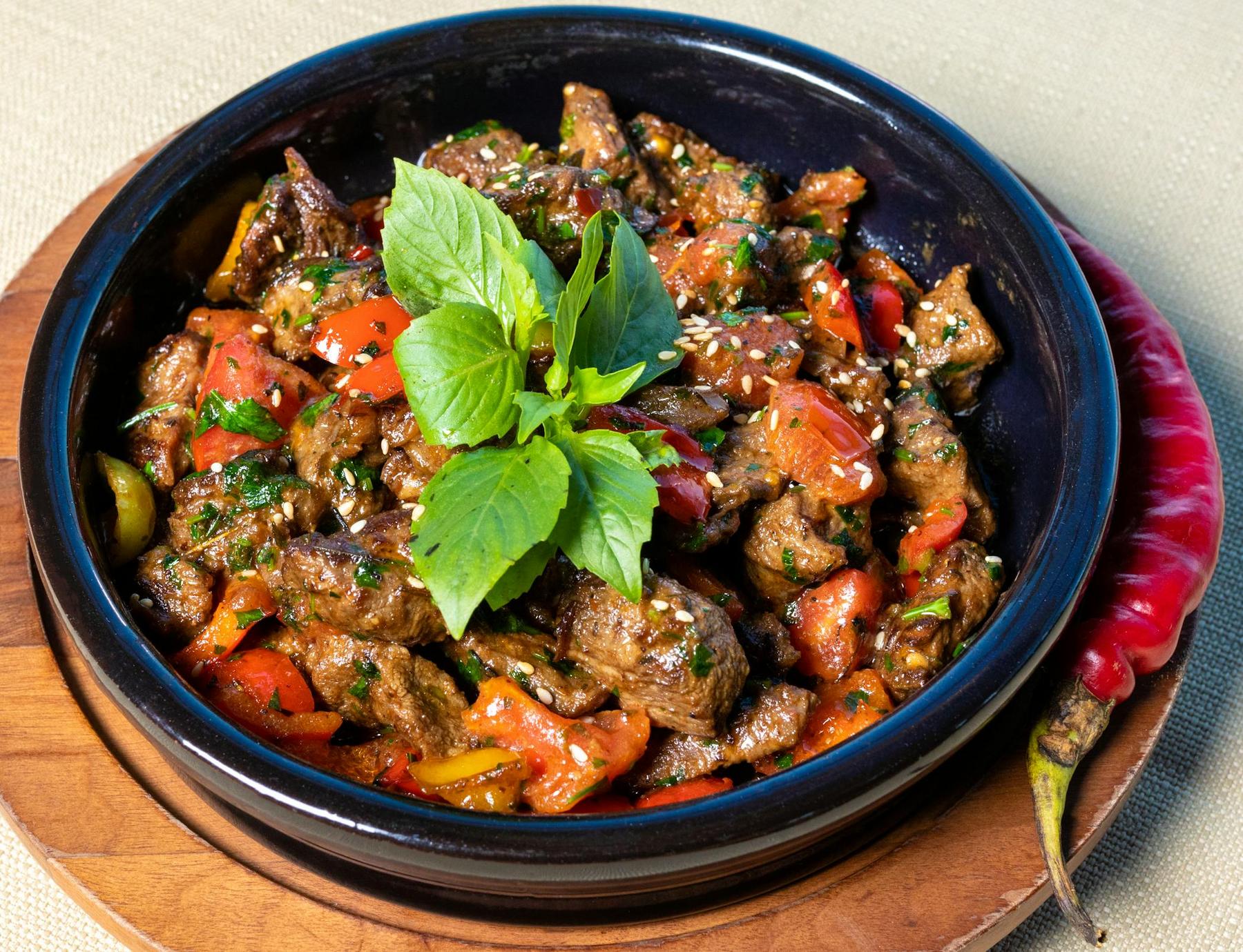 A black bowl filled with stir-fried beef, sliced red peppers, herbs, and sesame seeds, garnished with fresh basil, sits on a wooden platter next to a whole red chili pepper.