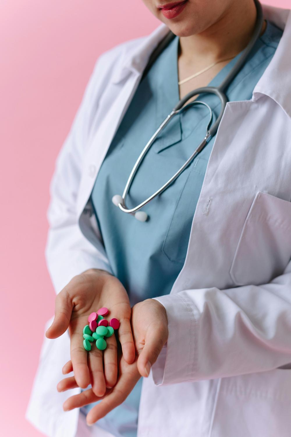 A healthcare professional in a lab coat and scrubs holds various colorful pills in their hands against a pink background.