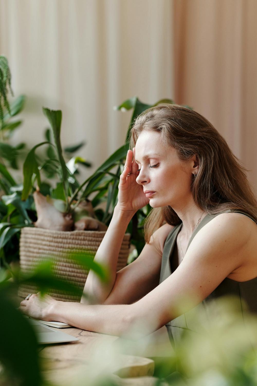 A woman sits at a table indoors with her hand to her temple, appearing deep in thought or focused, surrounded by green plants.