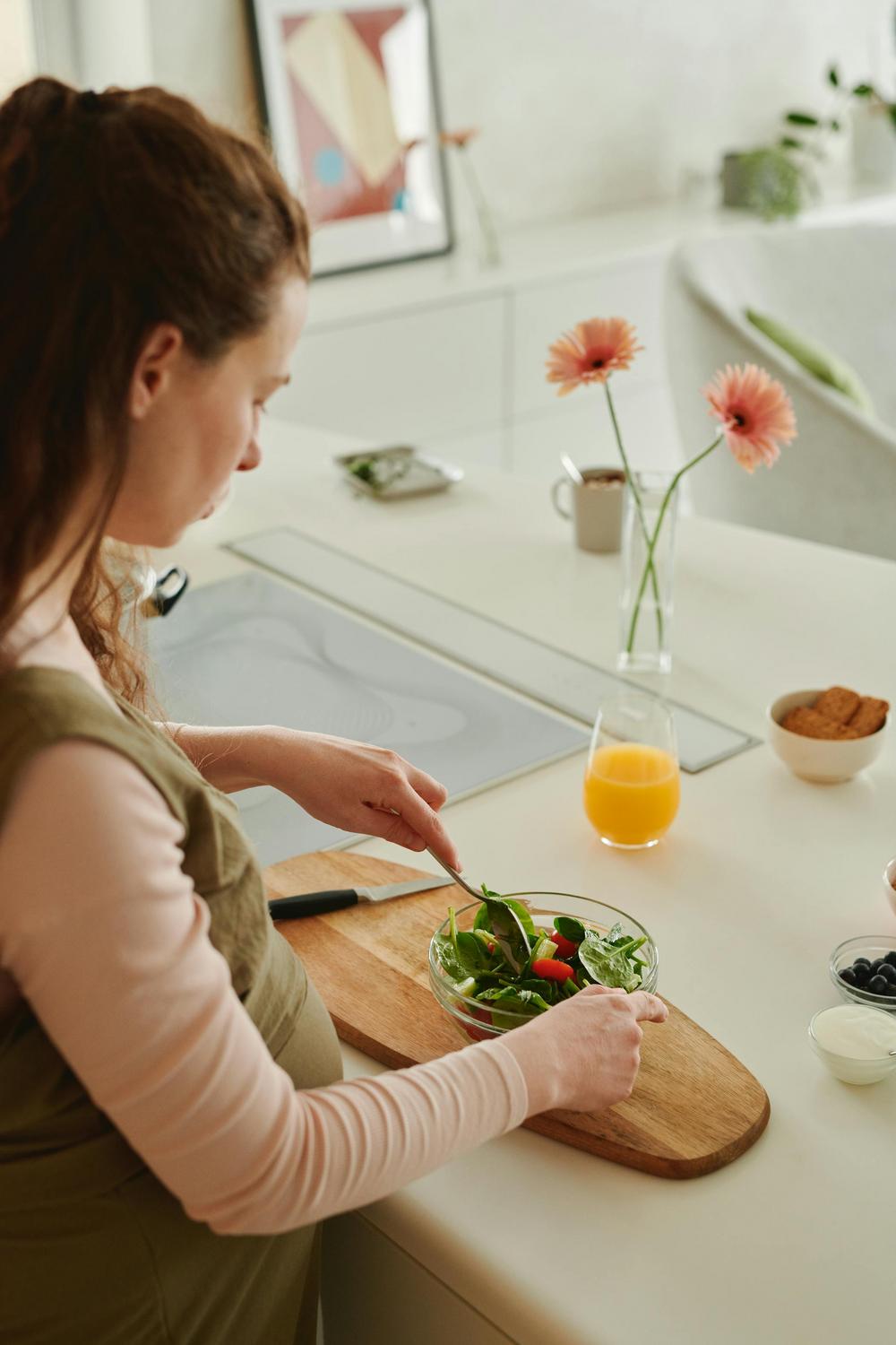 A person stands at a kitchen counter preparing a salad in a glass bowl. On the counter are a vase with flowers, a glass of orange juice, and small bowls with ingredients.