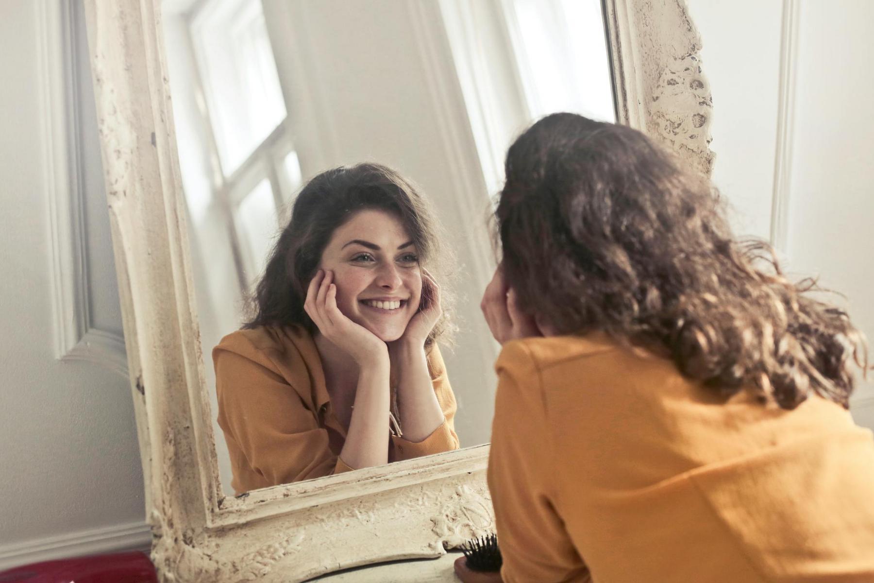 A woman with curly brown hair and a yellow top smiles and holds her face while looking at herself in a large ornate mirror.