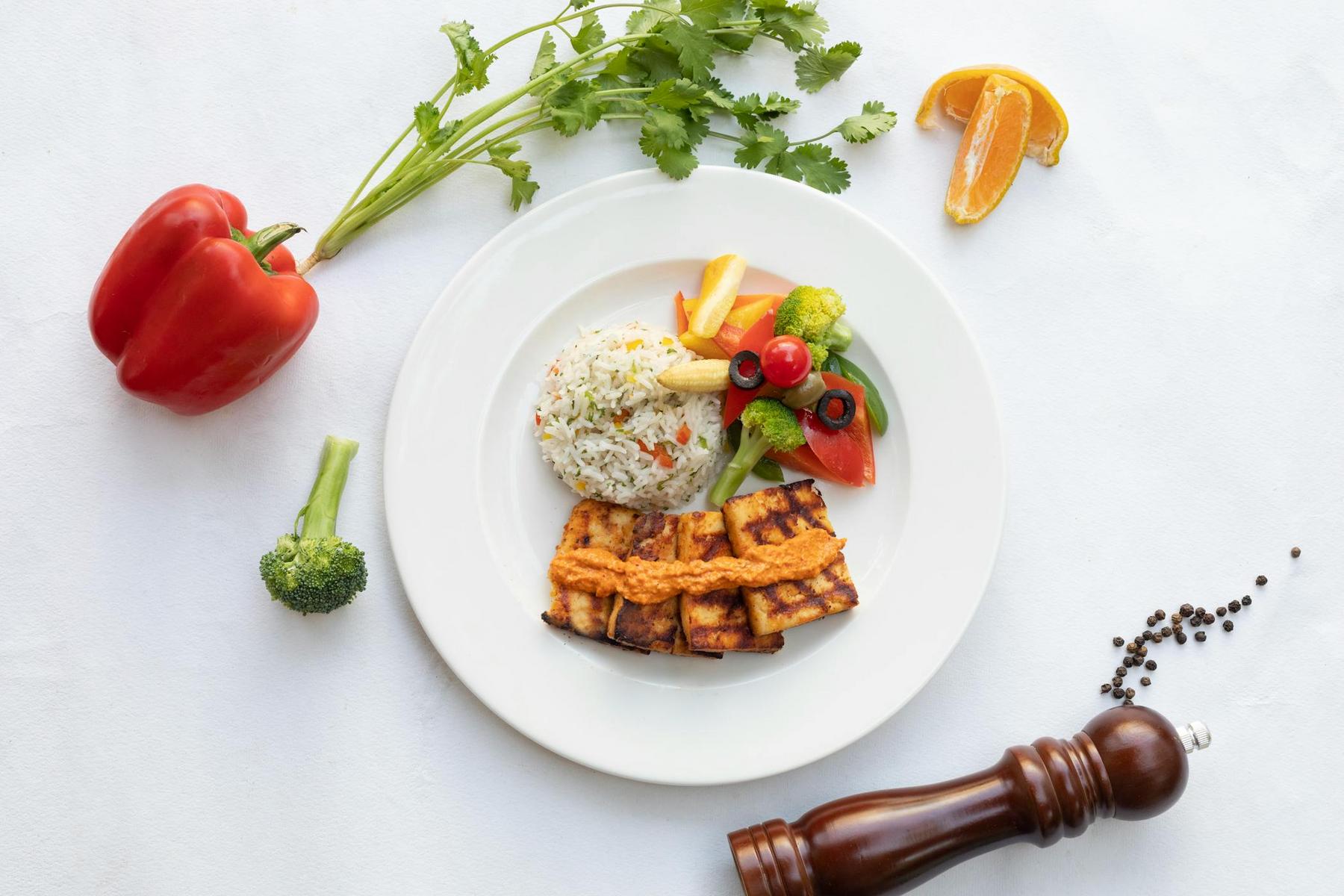 A white plate with grilled tofu, rice, and assorted vegetables sits on a white table with a red bell pepper, broccoli, cilantro, orange slice, pepper mill, and peppercorns nearby.
