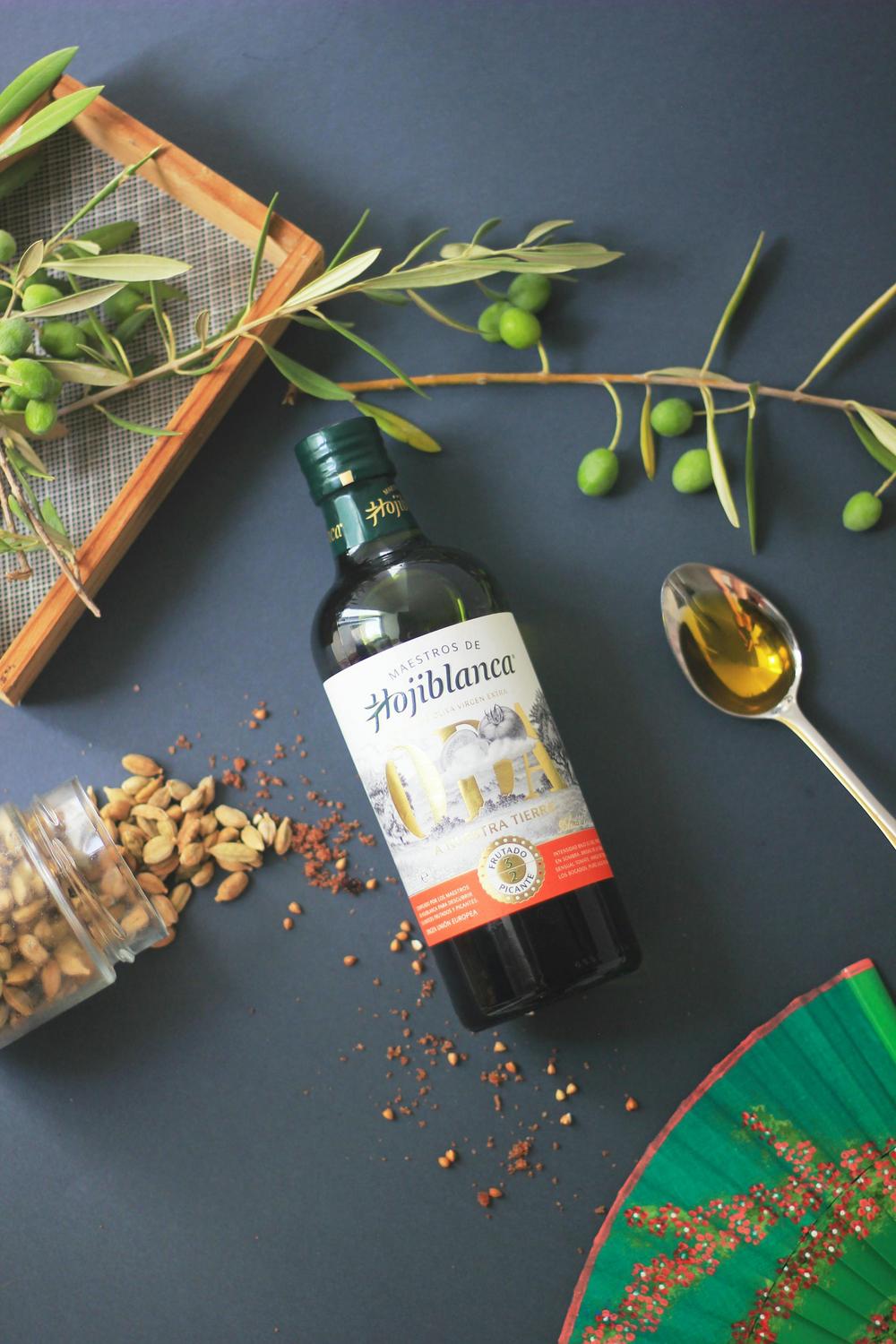 A bottle of Hojiblanca olive oil lies on a dark surface surrounded by green olives, cardamom pods, a spoon of oil, and scattered spices.