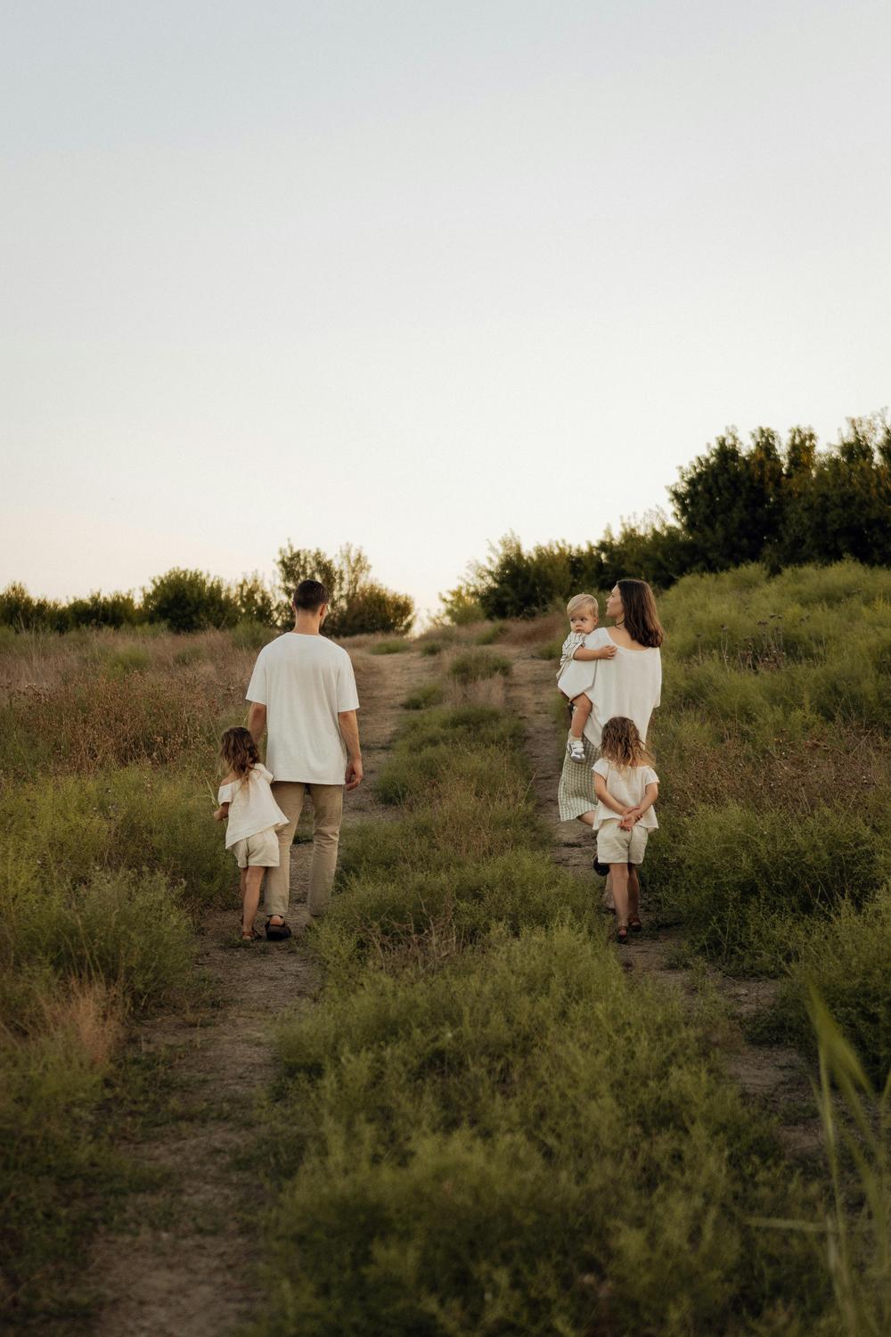 A family of five, dressed in light-colored clothing, walks up a grassy path outdoors, surrounded by greenery and under a pale sky.