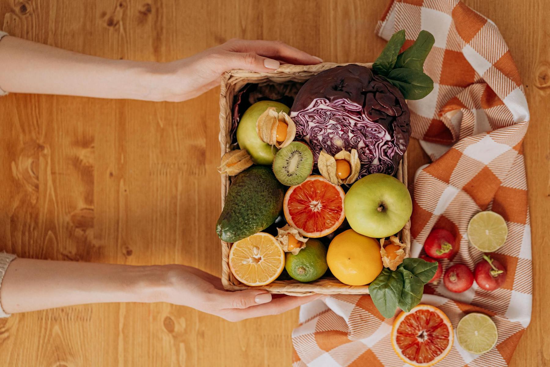 Hands holding a basket with red cabbage, avocado, citrus fruits, apples, kiwi, and greens on a wooden table with a checkered cloth.