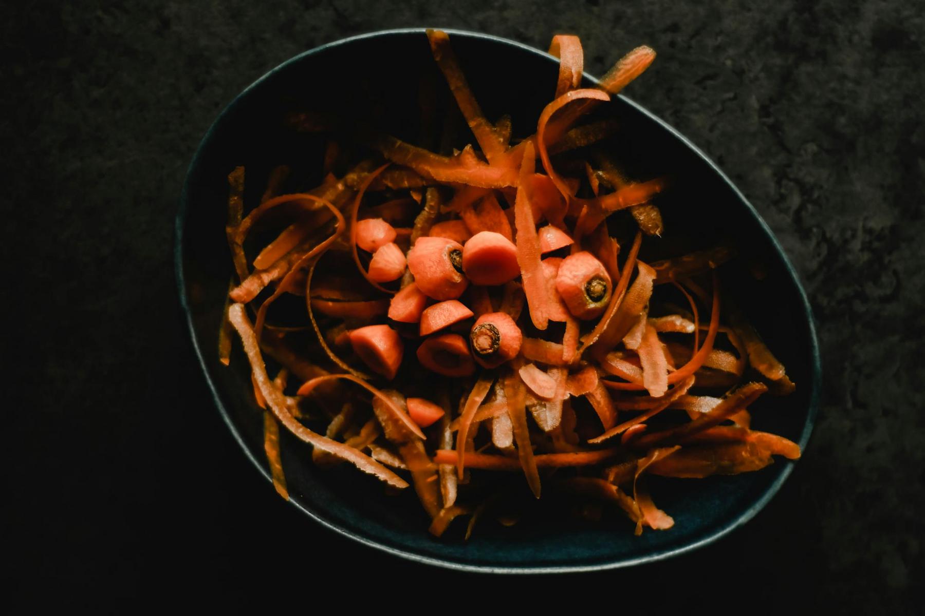 A black bowl containing carrot peels and sliced carrot pieces on a dark surface.