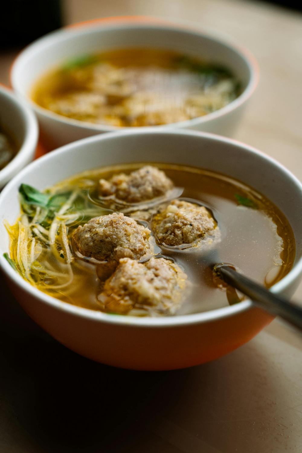 A close-up of a bowl of clear soup with meatballs, noodles, and leafy greens, with a spoon resting inside. Another similar bowl is visible in the background.