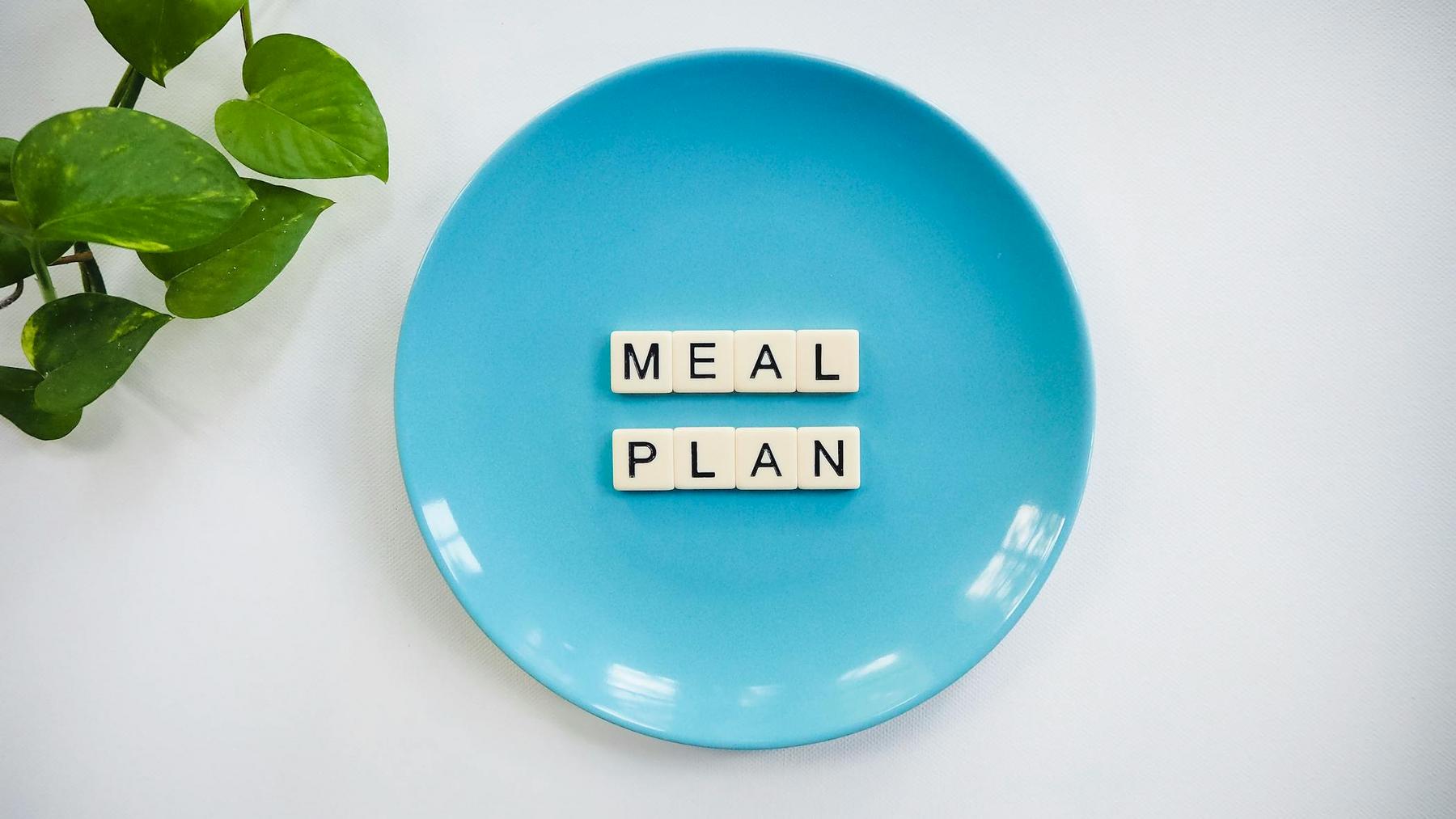 A blue plate with letter tiles spelling "MEAL PLAN" is placed on a white surface next to a green leafy plant.