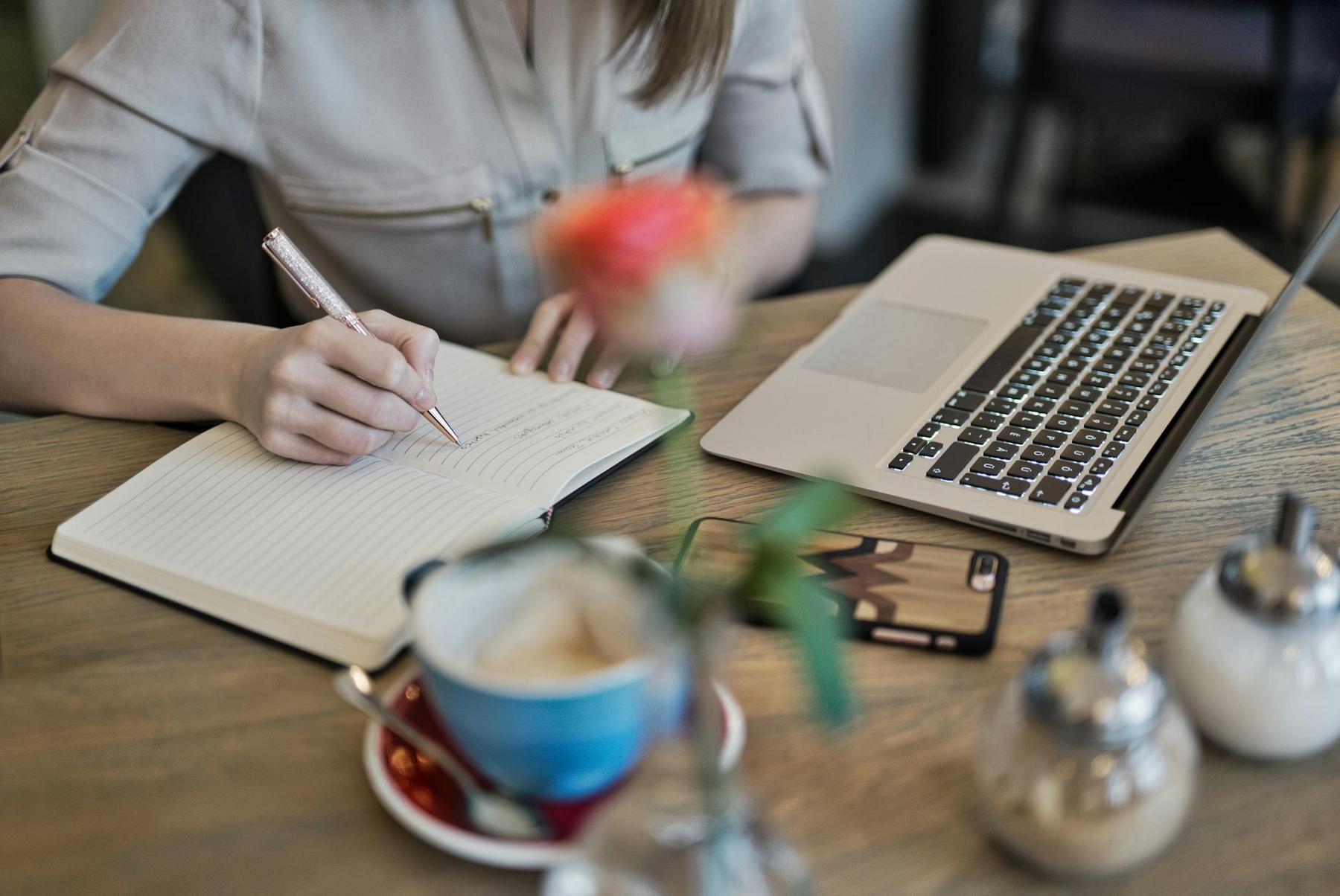Person writing in a notebook at a wooden table with a laptop, smartphone, coffee cup, and sugar containers nearby; a blurred flower is in the foreground.
