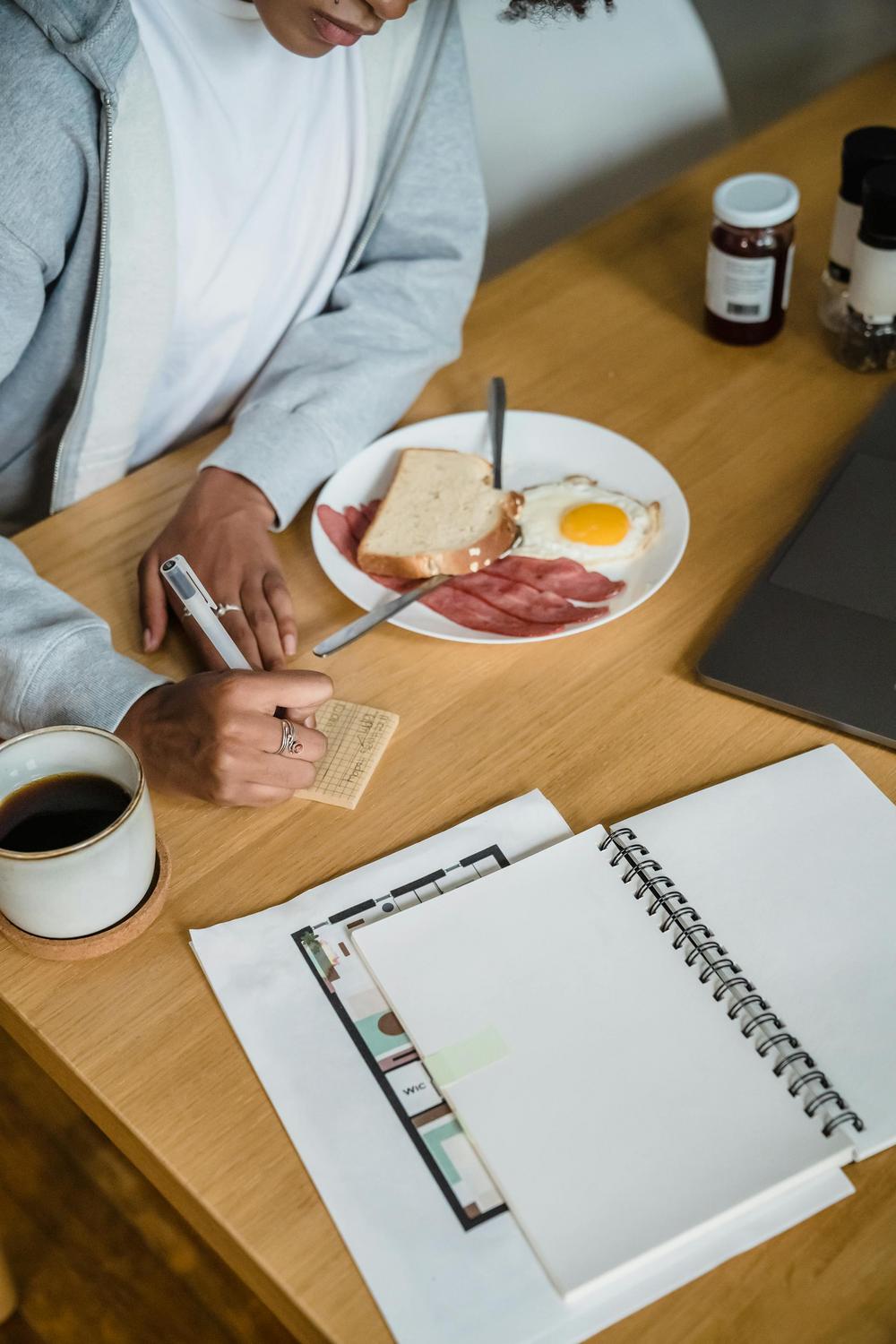 Person writing notes at a wooden table with a breakfast plate of toast, ham, and a fried egg, a cup of coffee, an open notebook, and a laptop.