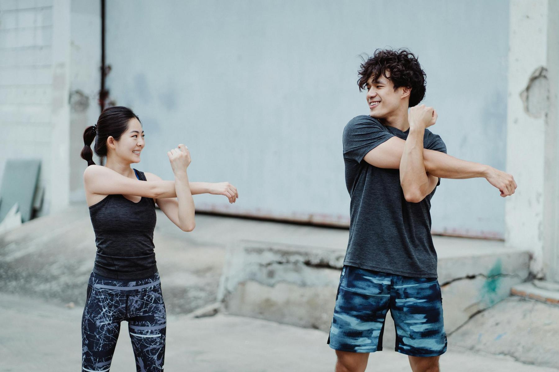 Two people in sportswear stretch their arms outdoors, facing each other and smiling, in front of a concrete building.