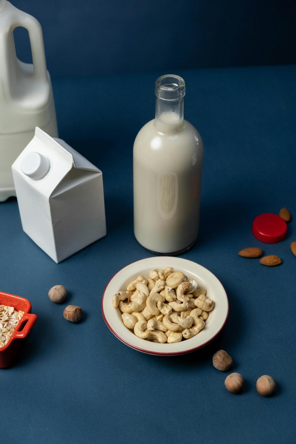 A bowl of cashews, a glass bottle of milk, a carton, a plastic jug, and scattered nuts are arranged on a blue surface.