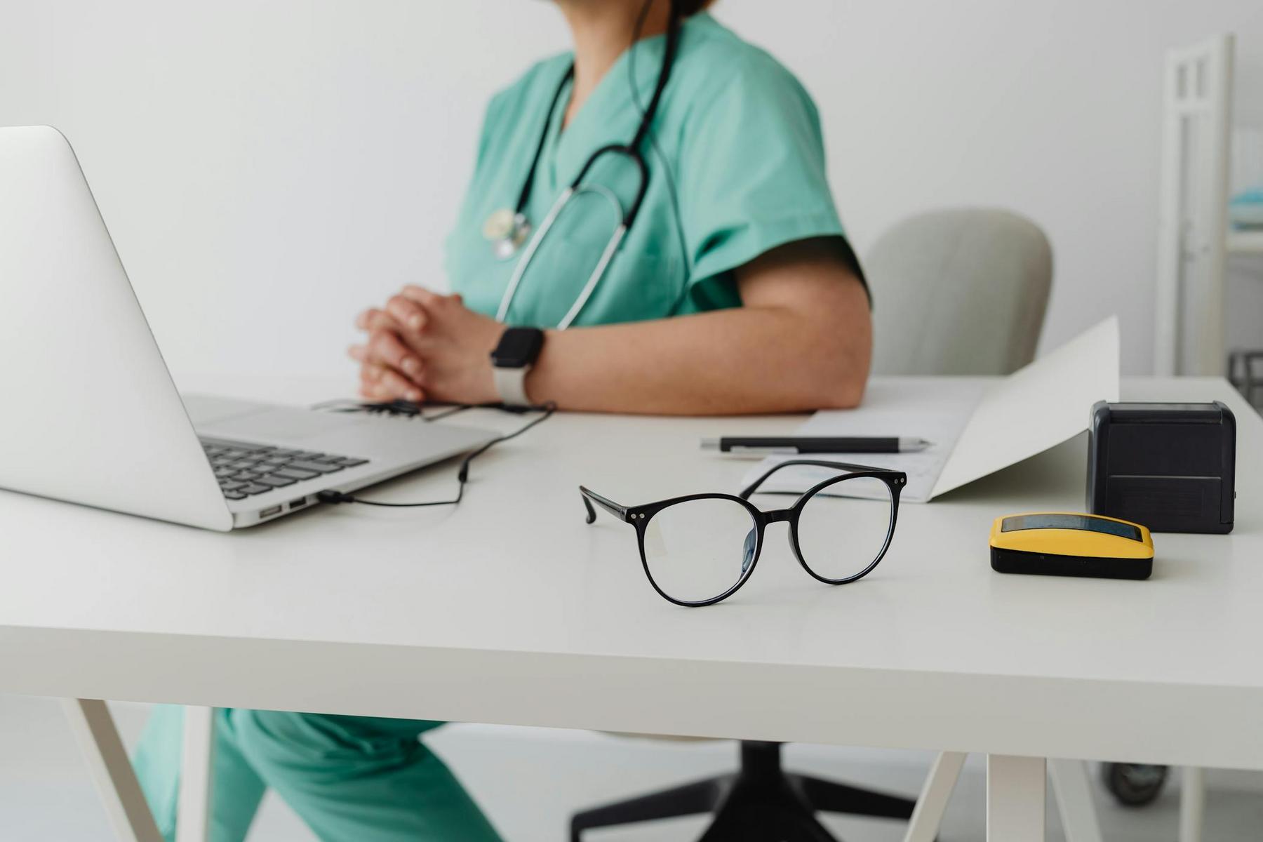 A person in medical scrubs sits at a desk with a laptop, paperwork, glasses, and a stethoscope visible.
