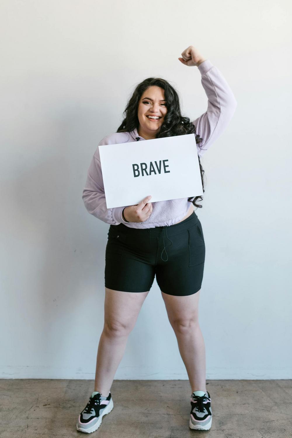 A woman stands smiling, holding a sign that says "BRAVE" and raising one fist in the air.