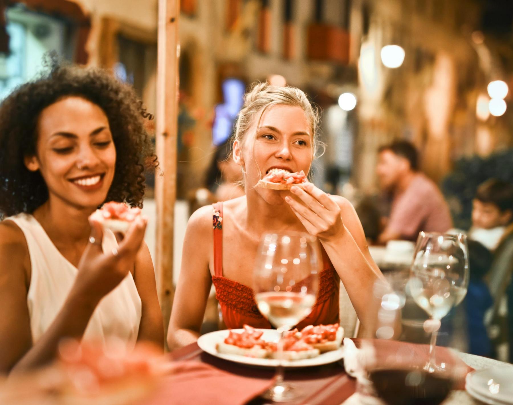 Two women are sitting at a restaurant table, eating food and smiling. Several wine glasses and plates with food are visible. The background shows other diners and warm lighting.