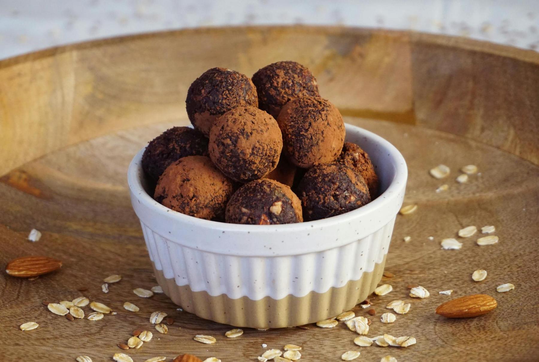 A small ceramic bowl filled with chocolate truffles, dusted with cocoa powder, placed on a wooden tray with scattered oats and almonds.