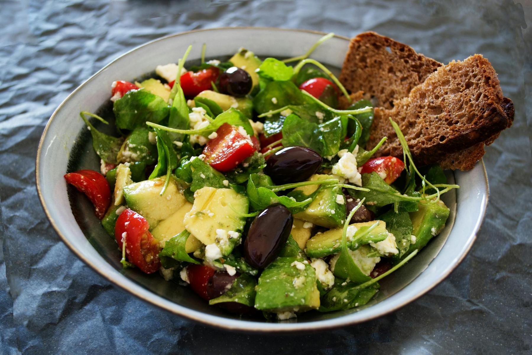A bowl of salad with avocado, cherry tomatoes, feta cheese, olives, leafy greens, and two slices of brown bread on the side.