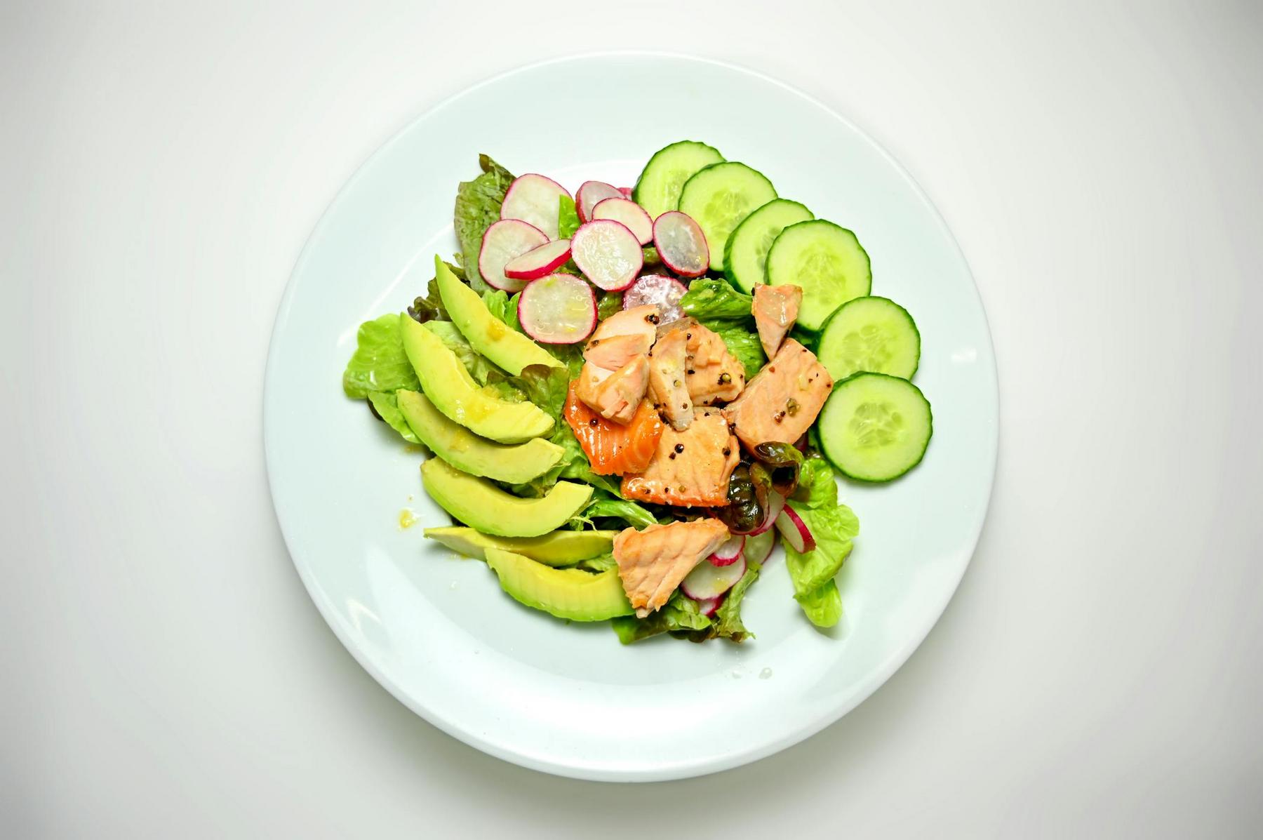 A white plate with sliced avocado, cucumber, radish, grilled salmon pieces, and mixed greens arranged neatly.