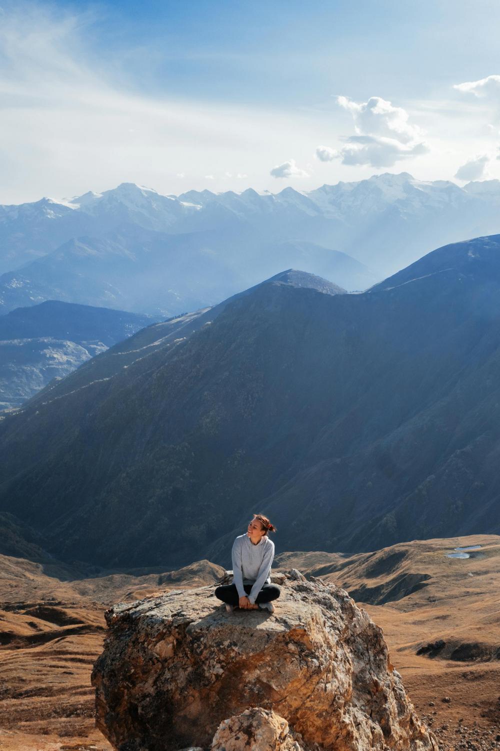 A person sits cross-legged on a large rock with mountains and a partly cloudy sky in the background.