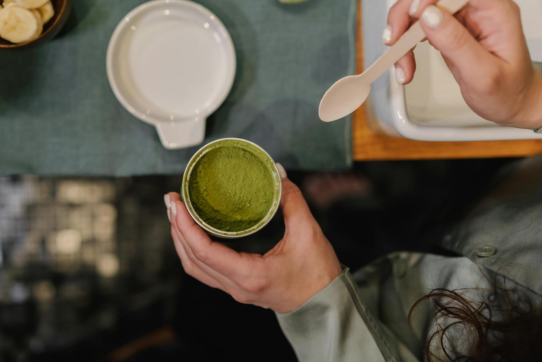 A person holds a container of green matcha powder in one hand and a wooden spoon in the other, with a white bowl and banana slices on a table nearby.