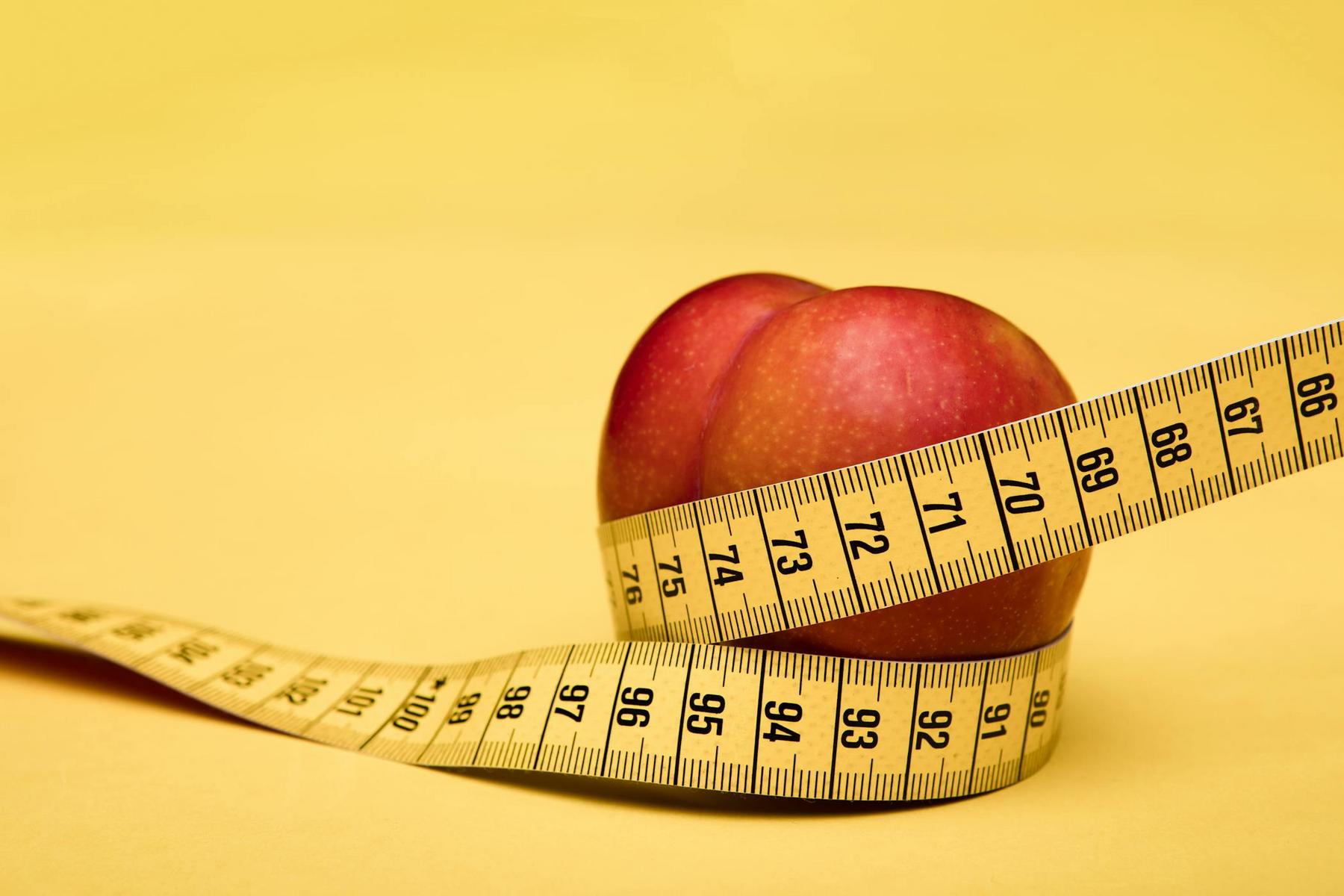 A red apple with a yellow measuring tape wrapped around it, set against a yellow background.