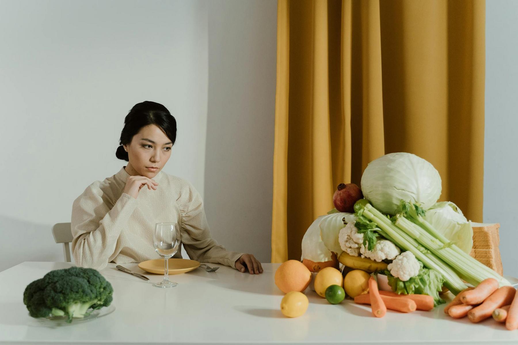 A woman sits at a table with an empty plate and glass, facing a large pile of assorted vegetables and fruits including cabbage, carrots, and broccoli.