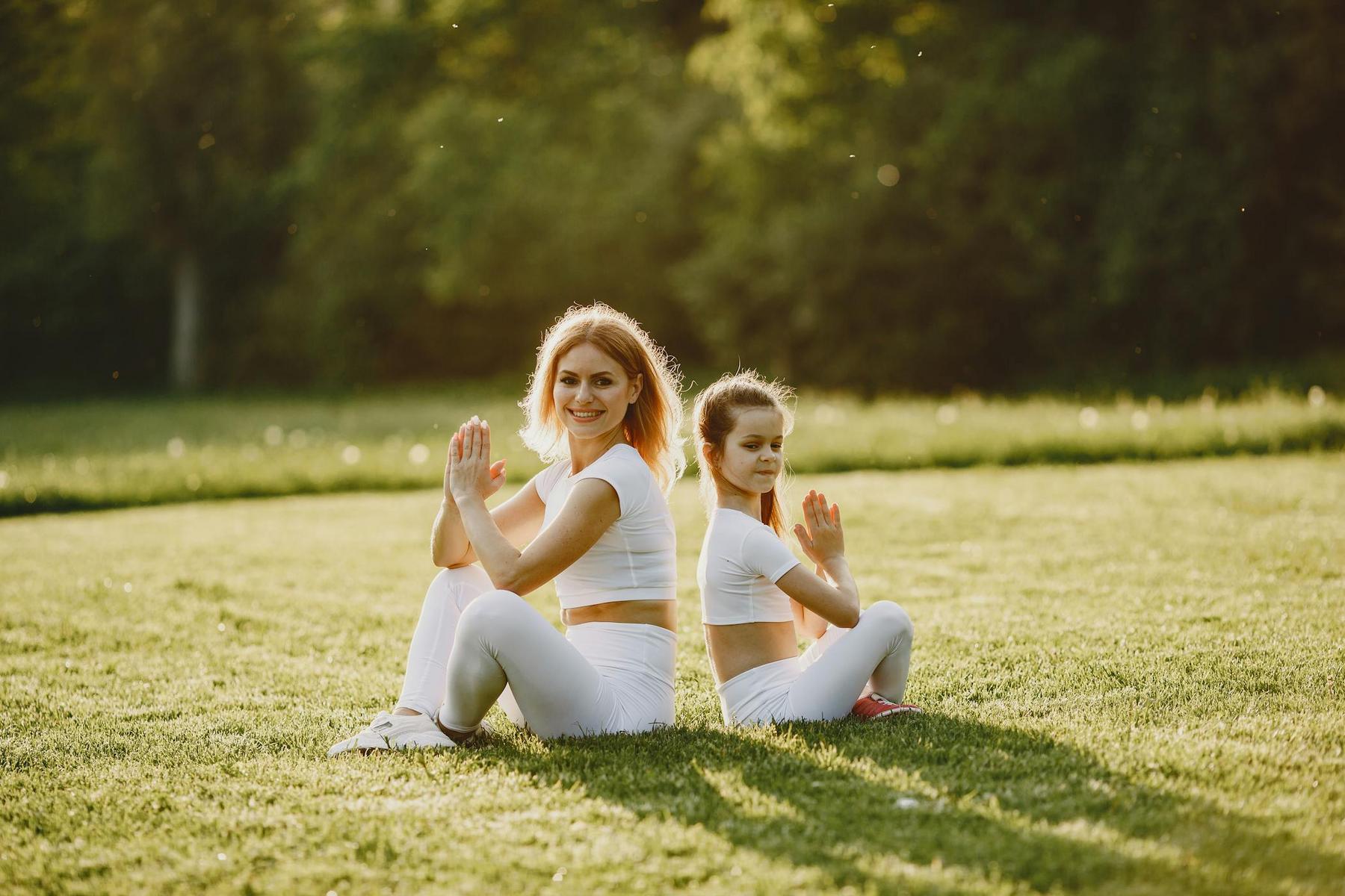 Two people wearing white athletic clothing sit back-to-back on grass in a yoga pose, with hands pressed together, outdoors in a sunlit park.