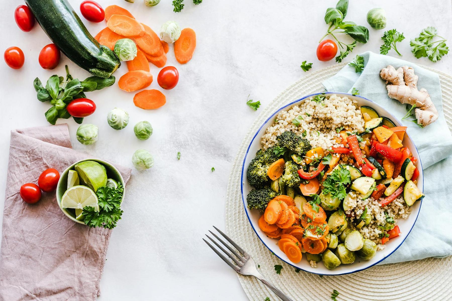 A bowl of quinoa topped with broccoli, carrots, bell peppers, and zucchini sits on a placemat, surrounded by fresh vegetables, lime wedges, and herbs on a white surface.