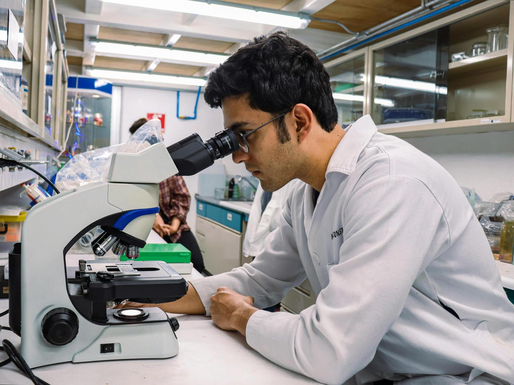A person wearing a lab coat observes a specimen through a microscope in a laboratory setting.