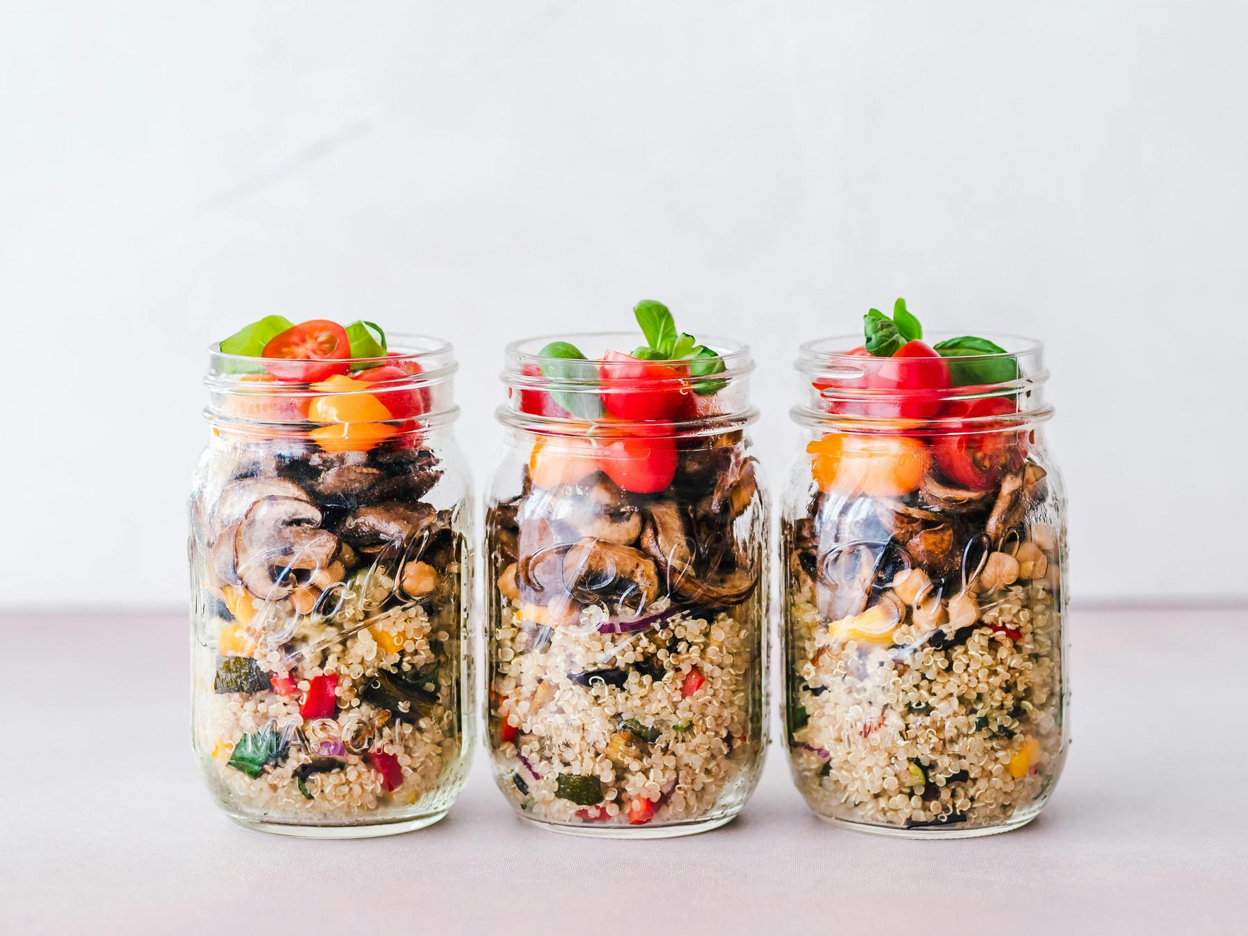 Three glass jars filled with layers of quinoa, roasted vegetables, chickpeas, and cherry tomatoes, topped with fresh basil leaves, arranged in a row on a light surface.