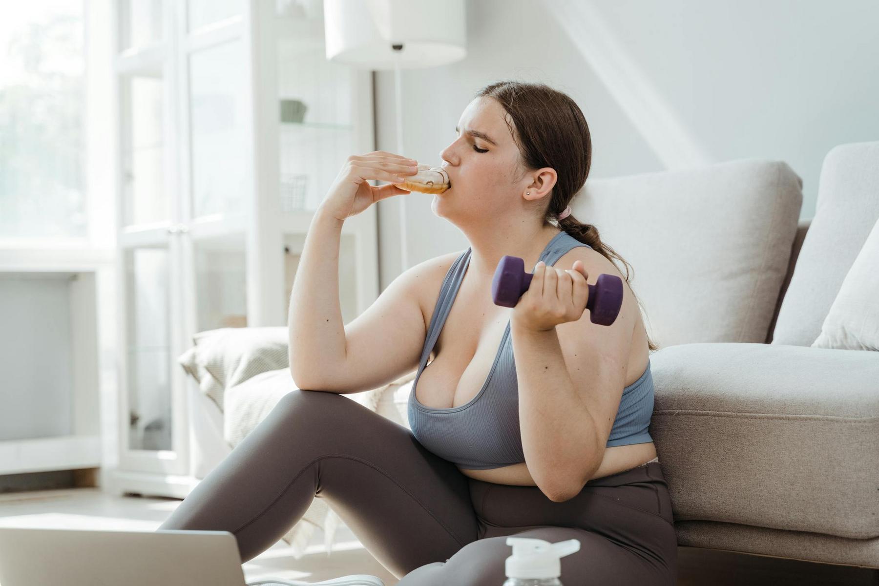 A person sits on the floor holding a purple dumbbell and eating a sandwich in a living room setting.