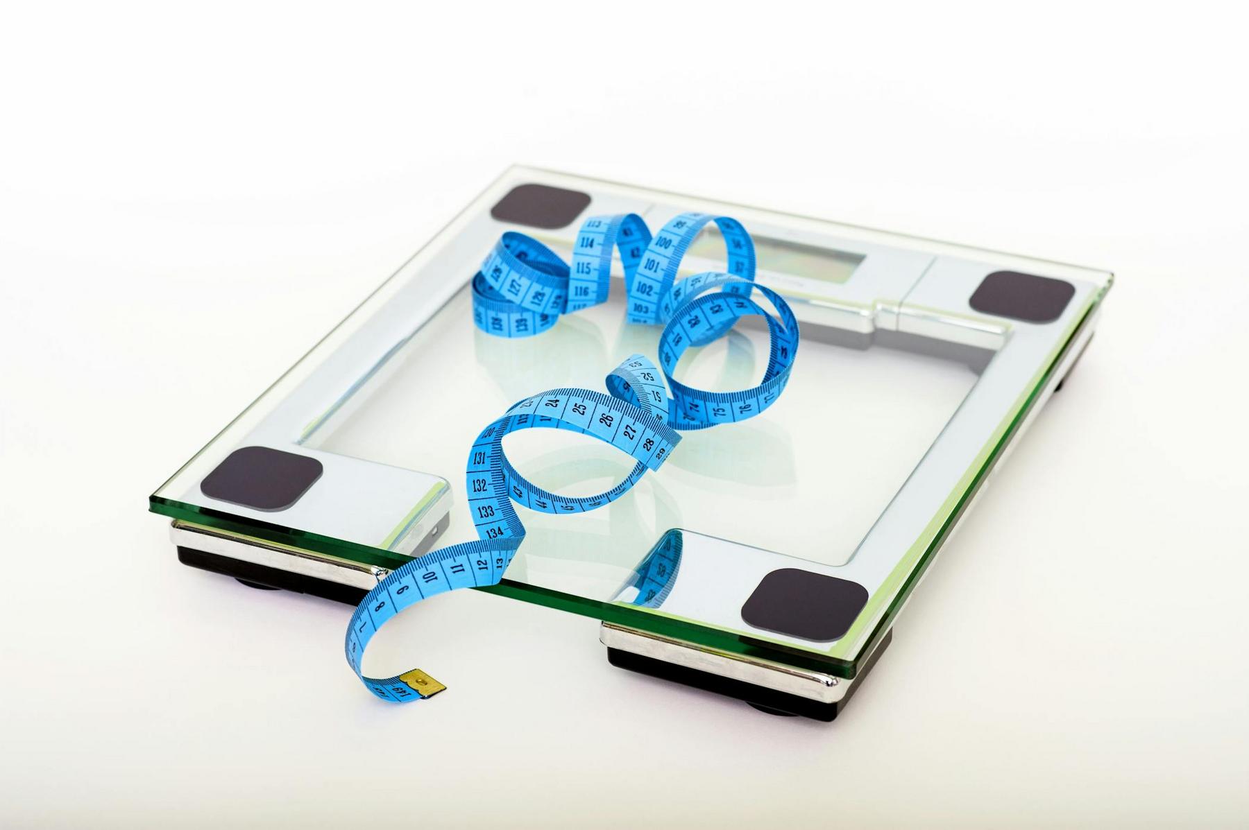 A glass digital bathroom scale with a blue measuring tape coiled on top, placed on a white surface.