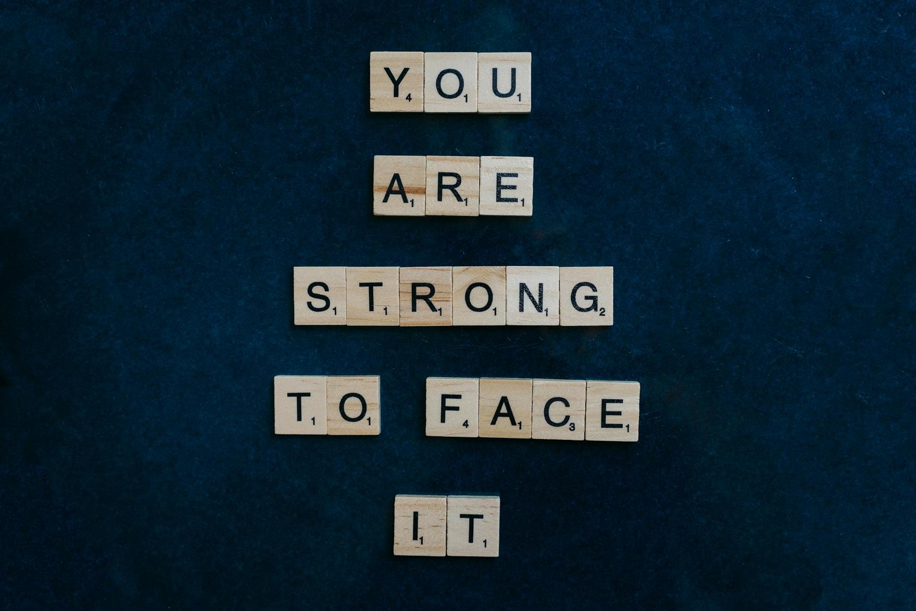 Wooden Scrabble tiles arranged on a dark surface spell out "You are strong to face it.