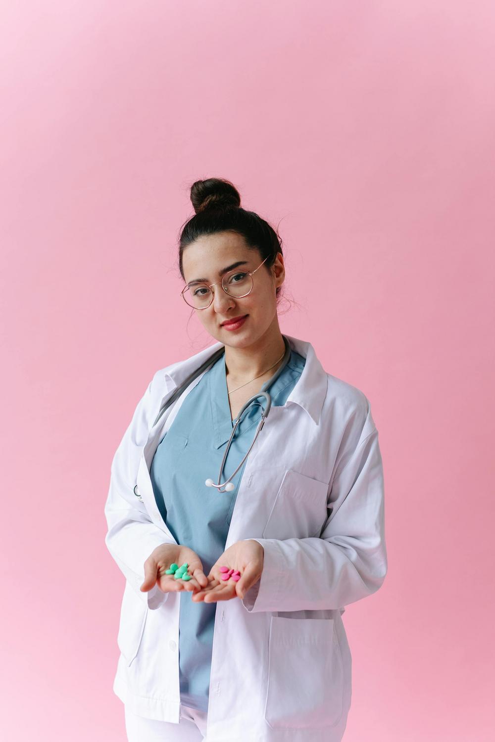 A medical professional wearing a lab coat and stethoscope holds green and pink pills in her hands, standing against a pink background.