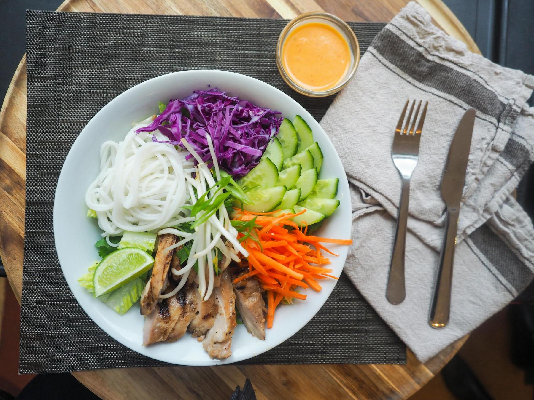 A bowl of grilled chicken, noodles, sliced cucumbers, red cabbage, carrots, lime wedge, and sprouts. A small jar of orange sauce is on the side. Cutlery on a gray napkin is placed nearby.
