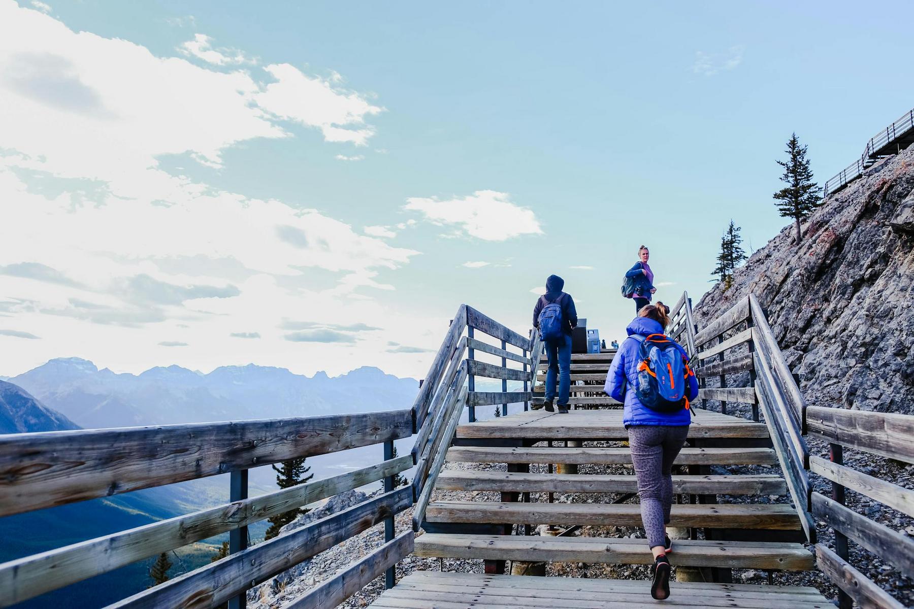 Three people with backpacks walk up wooden stairs on a mountain trail, surrounded by a wooden railing and distant mountain views under a partly cloudy sky.