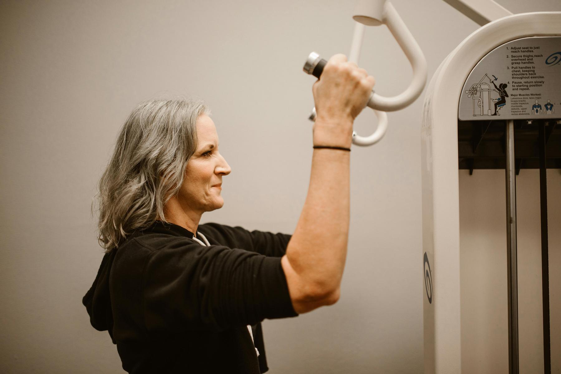 A woman with gray hair uses a seated weight machine, gripping the handles and lifting the weights in a gym setting.