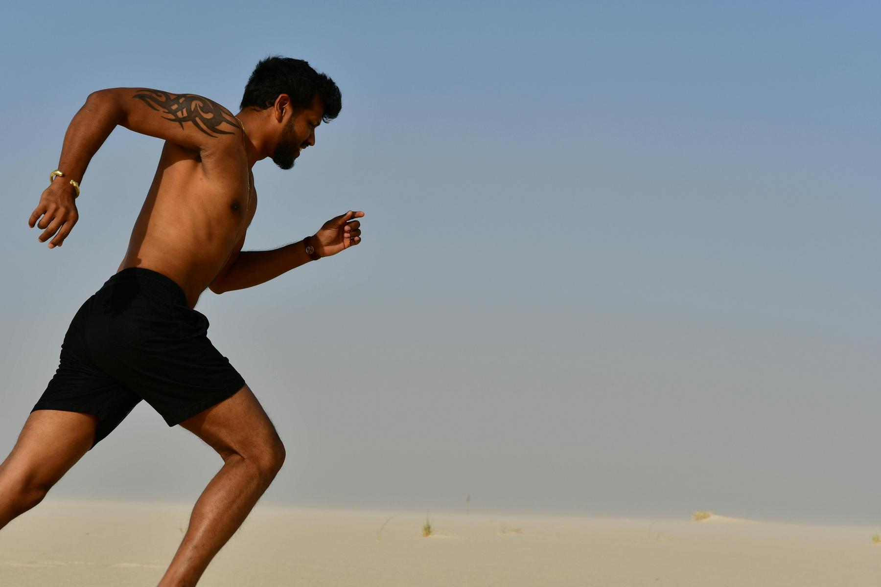 Shirtless man in black shorts with a tattoo on his arm running outdoors on sand under a clear sky.