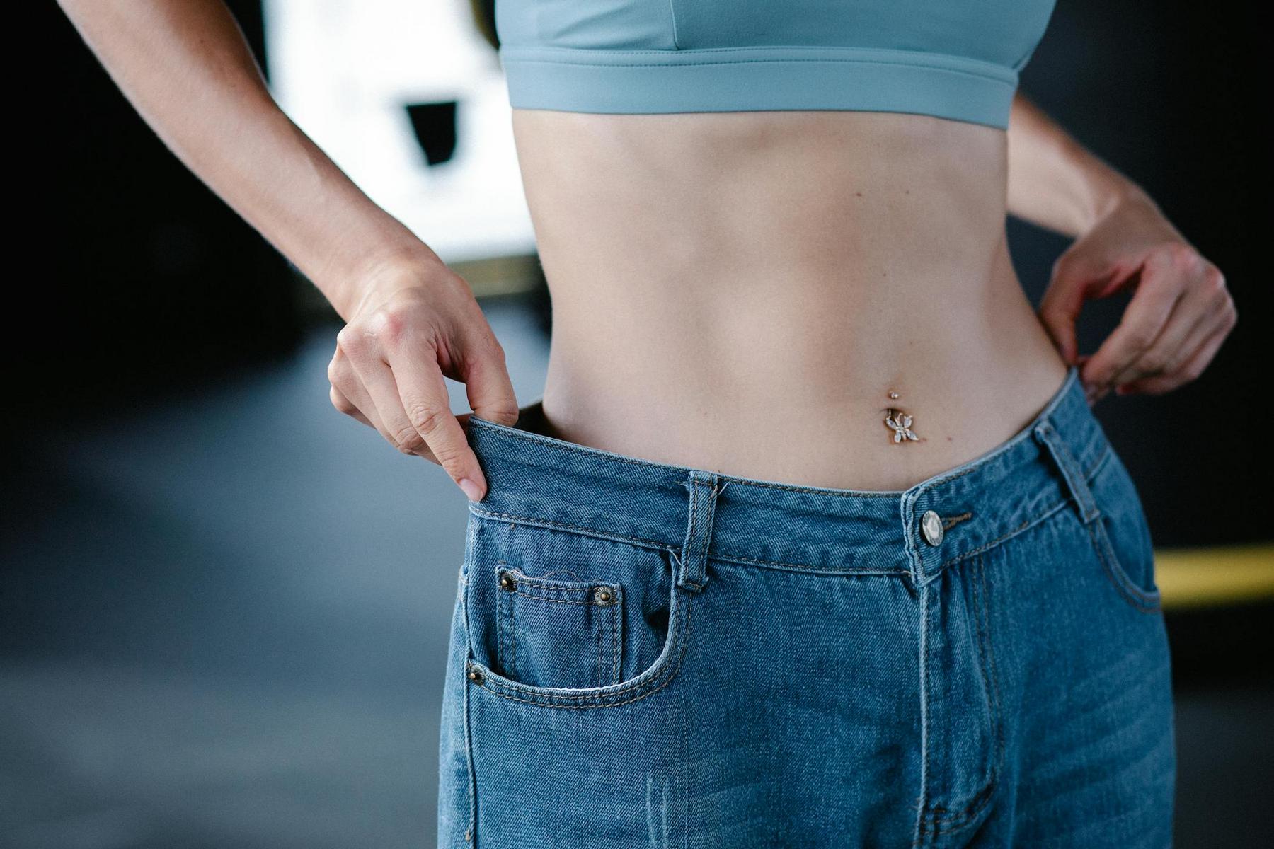 Person in a blue sports bra shows loose-fitting jeans by pulling the waistband away from their waist, with a belly button piercing visible.