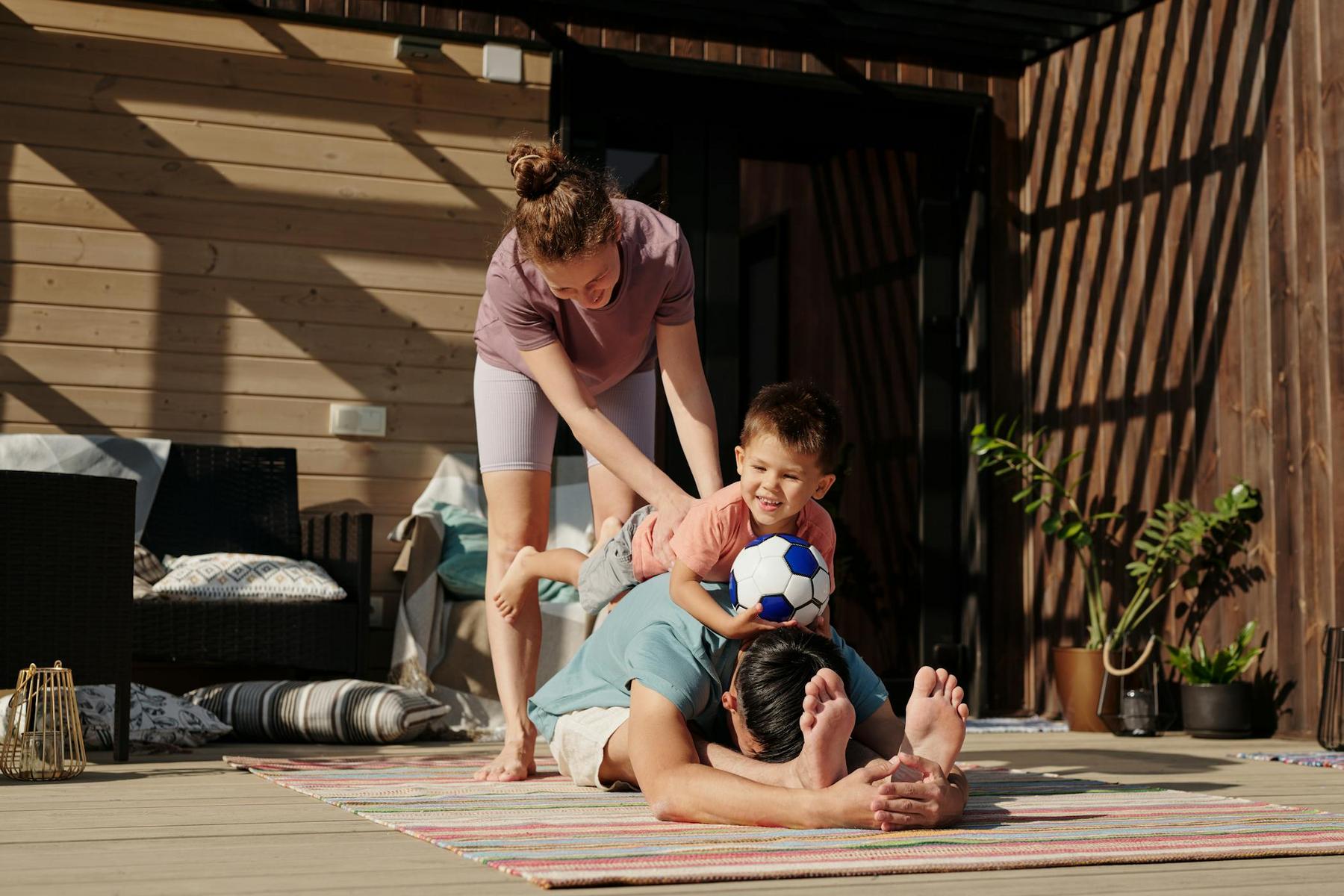 A child holding a small soccer ball lies on an adult’s back while another adult helps him balance, all on a striped mat outside a wooden house.