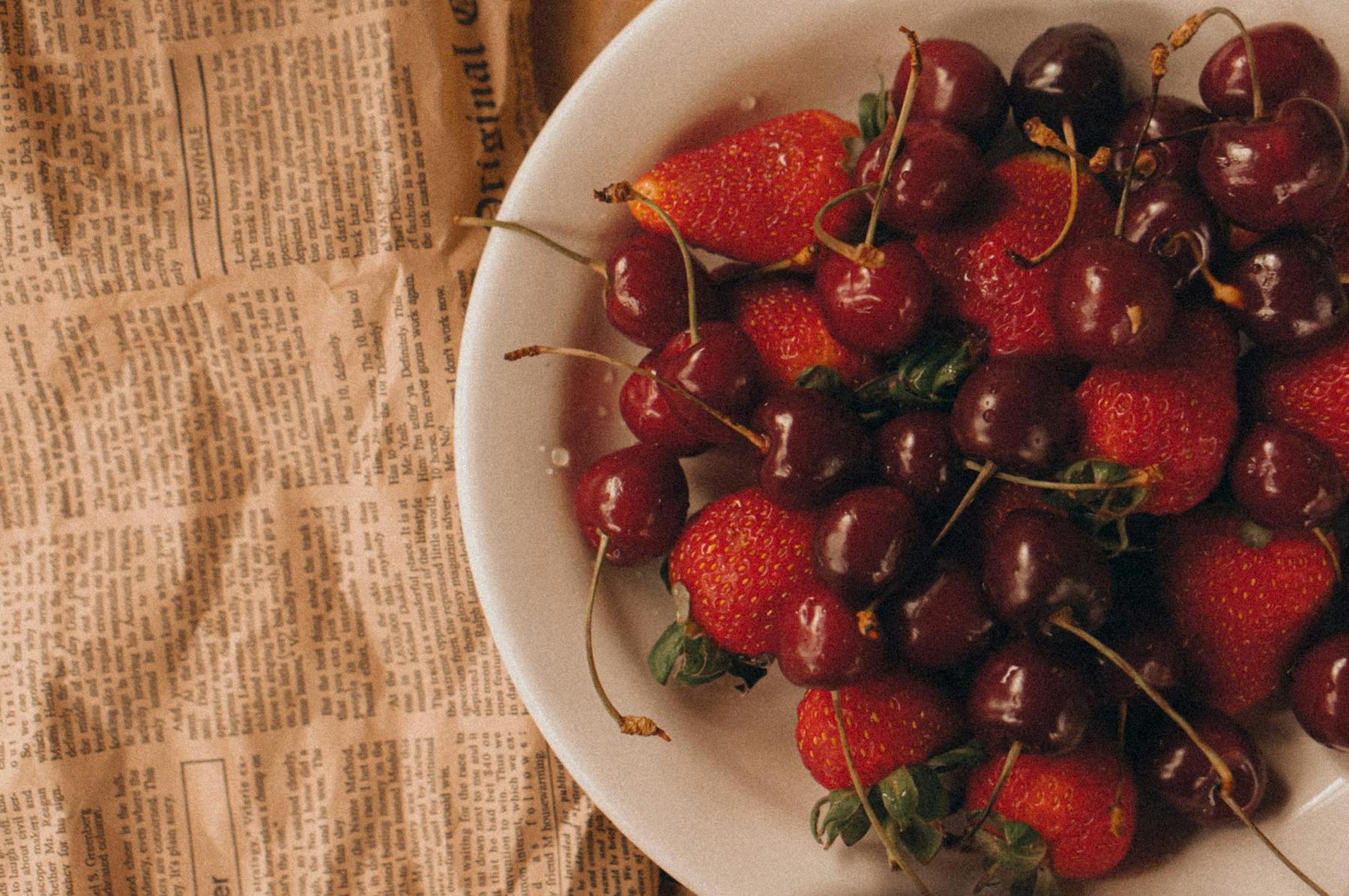 A white plate filled with fresh strawberries and cherries sits on a surface covered with old newspaper pages.