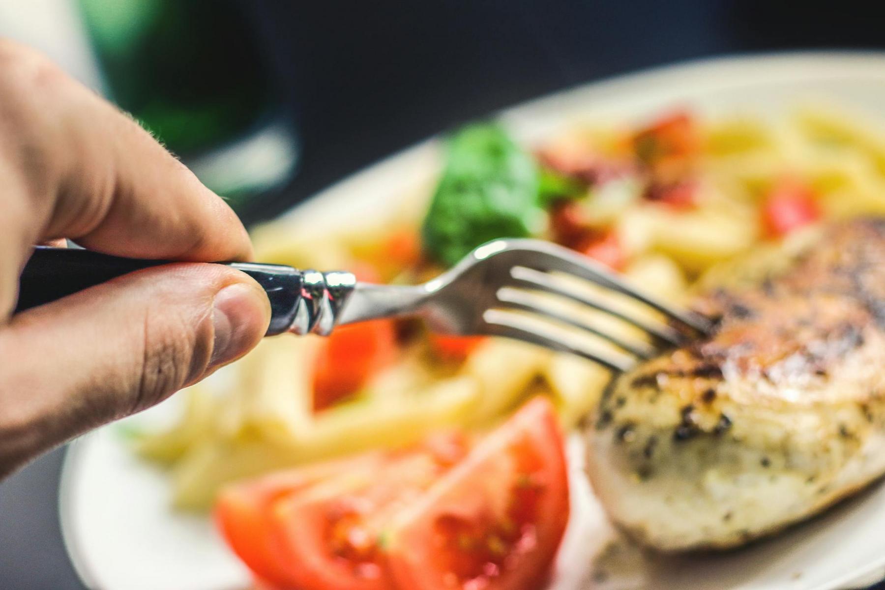 A person holding a fork prepares to eat grilled chicken, sliced tomatoes, pasta, and greens on a white plate.