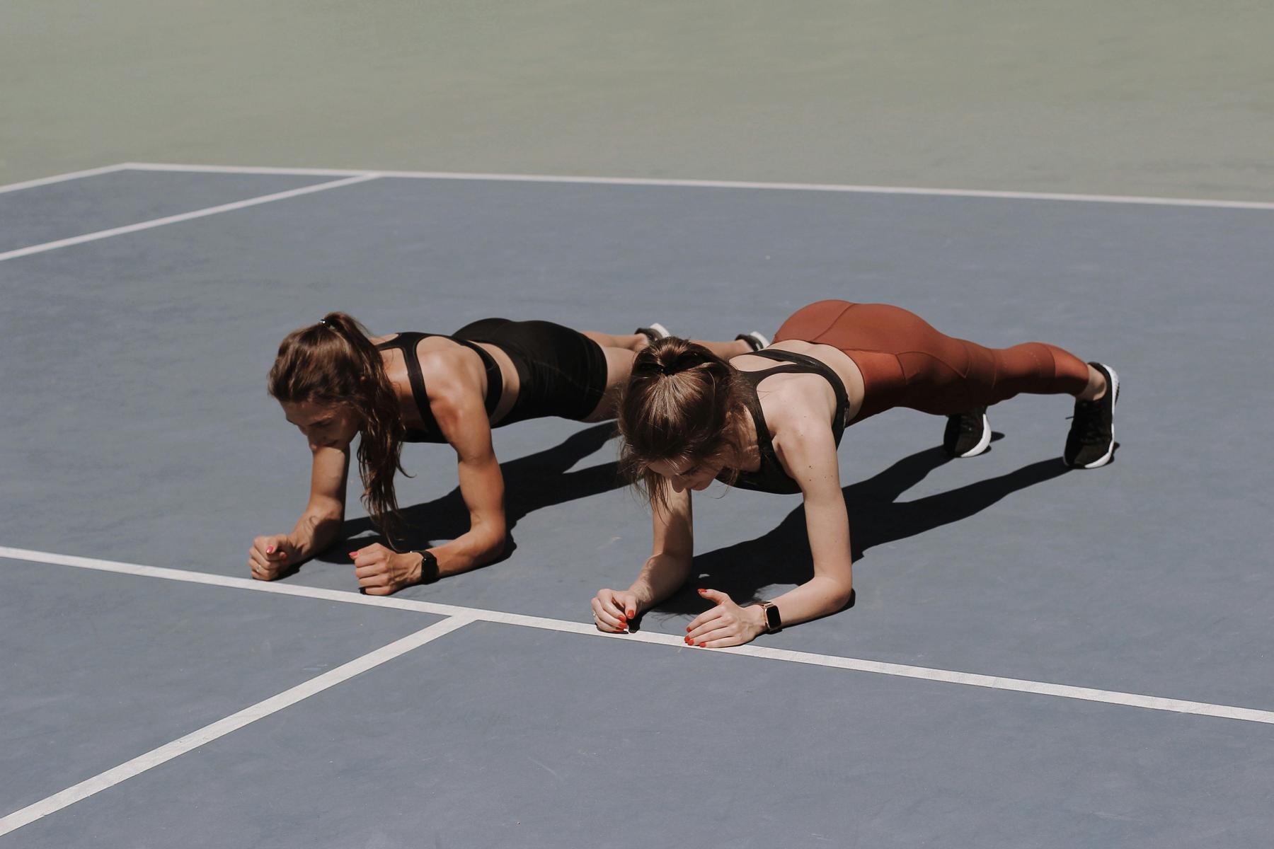 Two women in athletic wear perform plank exercises side by side on an outdoor sports court.