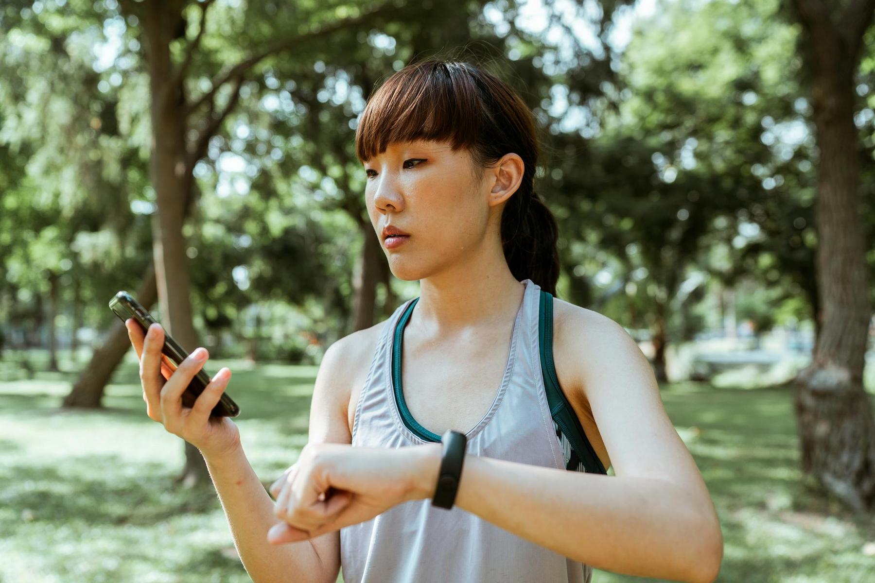 A woman in athletic wear checks her smartwatch and phone while standing outdoors in a park with trees in the background.