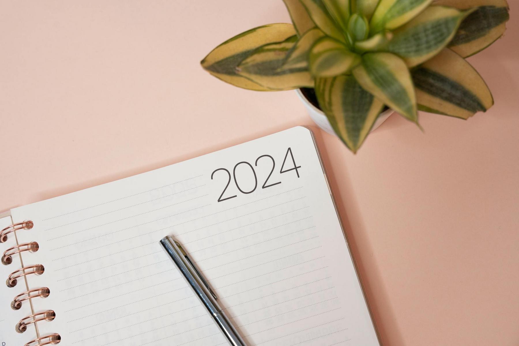 An open notebook with "2024" written at the top of the page, a pen resting on it, and a potted plant nearby on a light pink surface.