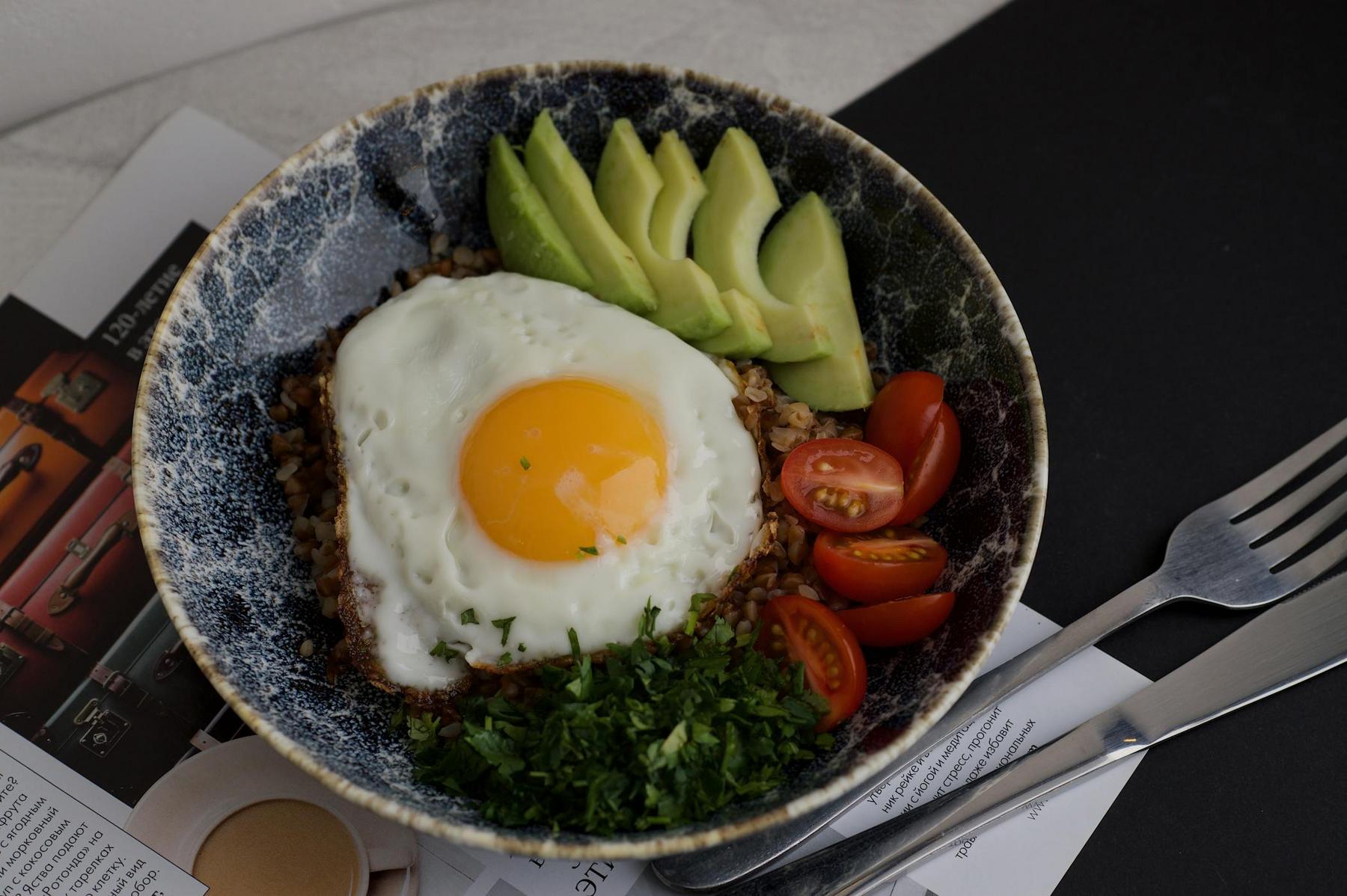 A bowl with a sunny-side-up egg on rice, sliced avocado, cherry tomatoes, and chopped herbs, placed on magazines with a fork and knife beside it.