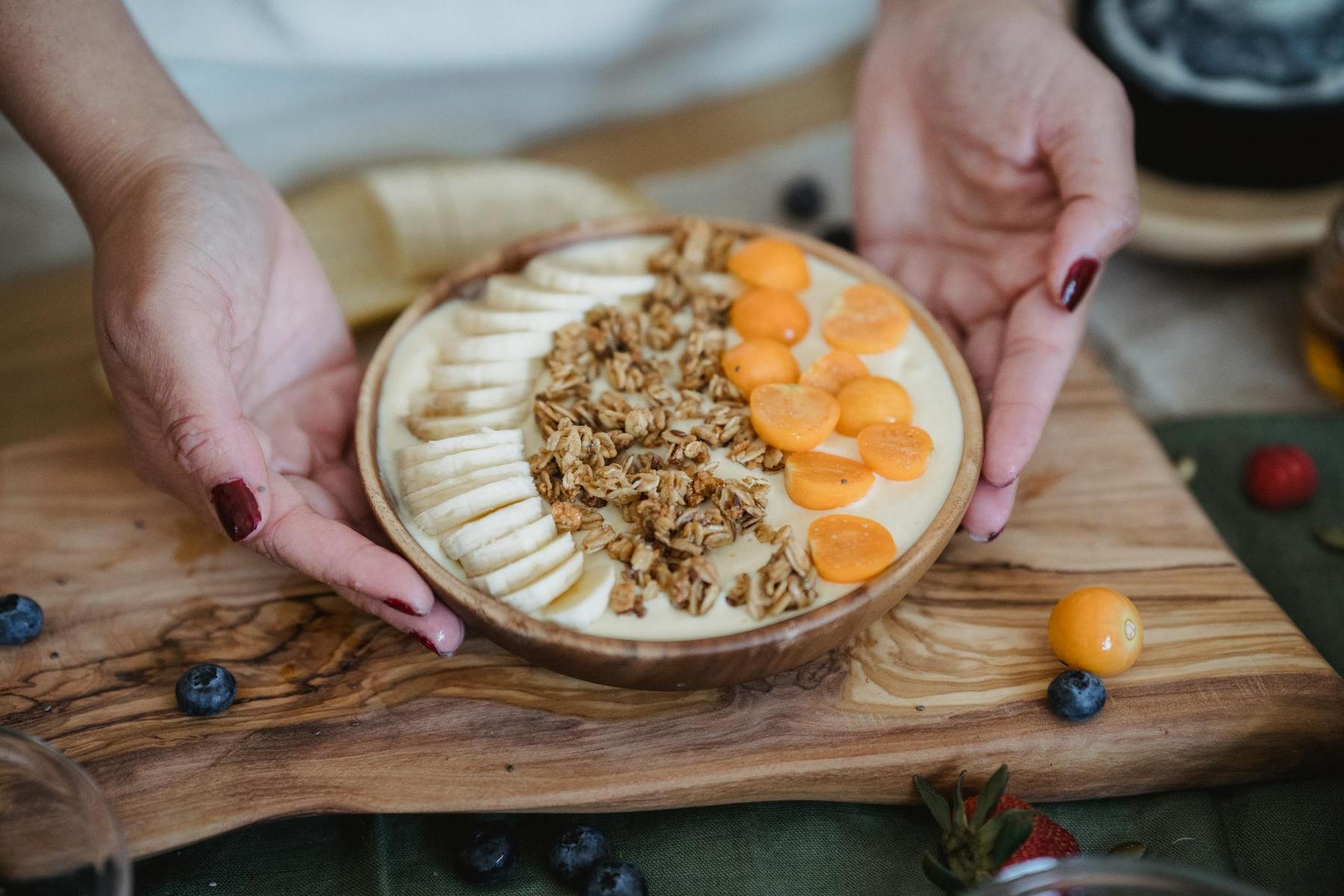 Person holding a smoothie bowl topped with sliced bananas, granola, and halved golden berries on a wooden board, surrounded by scattered blueberries and a strawberry.