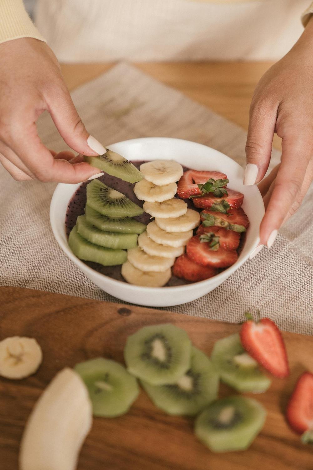 A person arranges sliced kiwi, banana, and strawberries on top of a smoothie bowl, with more fruit slices on a wooden board nearby.