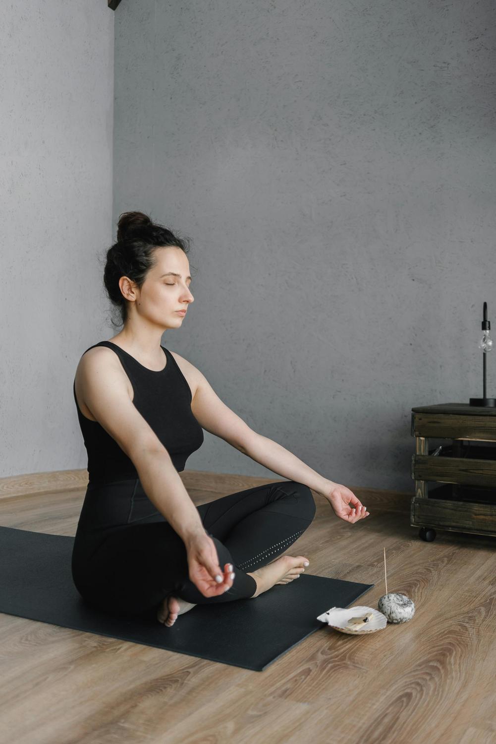 A woman in black athletic wear sits cross-legged on a yoga mat in a meditative pose, eyes closed, with incense and stones placed nearby on the floor.