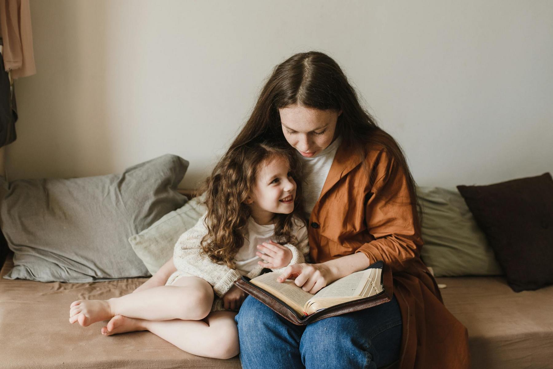 A woman sits on a couch reading a book to a smiling young girl who is leaning against her. Pillows are in the background.